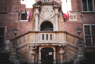 A historic brick building facade with an ornate stone staircase. Two classical statues support the entrance, surrounded by intricate carvings. Flags hang near a plaque on the wall, and traditional street lamps flank the stairway.