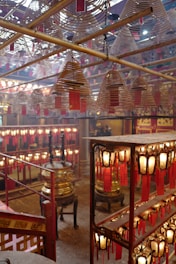 An interior setting with numerous spiral incense coils hanging from the ceiling, each with attached red tags. Soft, warm lighting emanates from surrounding lanterns that feature similar red tags, creating an inviting and peaceful atmosphere. In the background, large brass or gold-colored incense burners add to the traditional ambiance.