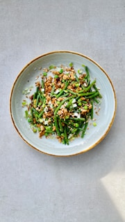 A ceramic bowl contains a visually appealing dish composed of green vegetables such as green beans and peas, mixed with small chunks of a brown protein source, garnished with sliced scallions and sprinkled with sesame seeds. The presentation is clean and minimalistic with a neutral background, allowing the vibrant colors of the dish to stand out.