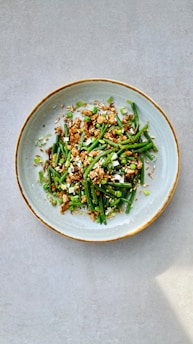 A ceramic bowl contains a visually appealing dish composed of green vegetables such as green beans and peas, mixed with small chunks of a brown protein source, garnished with sliced scallions and sprinkled with sesame seeds. The presentation is clean and minimalistic with a neutral background, allowing the vibrant colors of the dish to stand out.