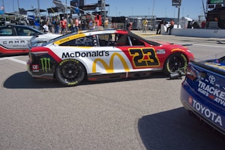 A brightly colored race car with the number 23 and prominent McDonald's and Monster Energy branding is parked in a pit area. The car features red, yellow, and white colors with Goodyear tires. Nearby are other vehicles and a group of people standing in the background, indicating a lively racing event setting.