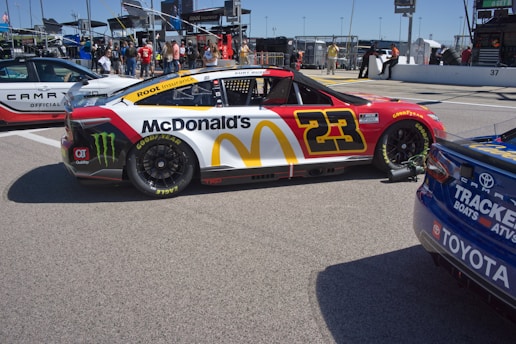 A brightly colored race car with the number 23 and prominent McDonald's and Monster Energy branding is parked in a pit area. The car features red, yellow, and white colors with Goodyear tires. Nearby are other vehicles and a group of people standing in the background, indicating a lively racing event setting.