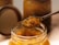 A jar filled with a chunky, golden vegetable mixture with seeds, possibly pickled or preserved, is opened. A spoon is lifting a portion of this mixture, showcasing its texture and ingredients. The background features more jars, suggesting a kitchen or food storage environment.