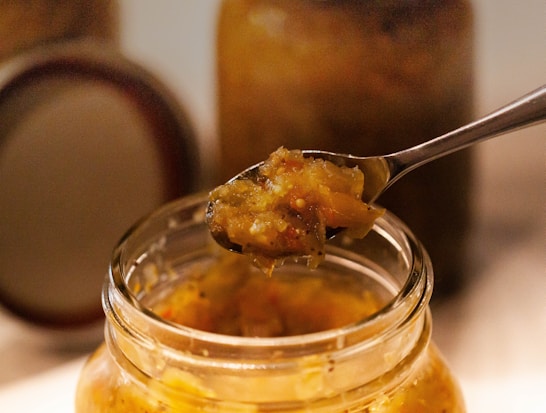 A jar filled with a chunky, golden vegetable mixture with seeds, possibly pickled or preserved, is opened. A spoon is lifting a portion of this mixture, showcasing its texture and ingredients. The background features more jars, suggesting a kitchen or food storage environment.