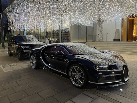 Two luxury cars, including a sports car and an SUV, are parked in front of a building adorned with shimmering decorative lights. The scene is set in an urban environment on a paved street, with the lighting creating a festive atmosphere.