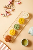 A visually appealing arrangement featuring a branch with pink blossoms, a set of colorful mooncakes on a transparent tray scattered with gold flakes, a white teapot, and a cup of green tea. The mooncakes are of varying colors and designs, each one intricately detailed and invitingly arranged.