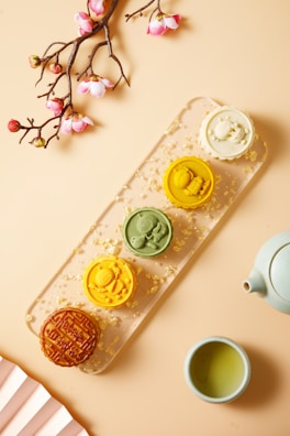 A visually appealing arrangement featuring a branch with pink blossoms, a set of colorful mooncakes on a transparent tray scattered with gold flakes, a white teapot, and a cup of green tea. The mooncakes are of varying colors and designs, each one intricately detailed and invitingly arranged.
