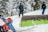 A skier in a bright blue and red outfit is navigating a snow-covered slope. The person appears to be in motion, with snow spraying around them. In the background, two other individuals dressed in winter gear are standing with skis, surrounded by snow-draped trees.