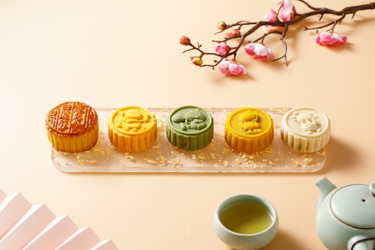 A tray holds five colorful mooncakes with intricate designs, arranged in a row. Nearby, there is a teapot and a cup filled with green tea. A branch with pink and white flowers decorates the scene, enhancing its aesthetic appeal.