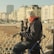 An elderly person wearing a white beret, pink scarf, and gloves is seated on a portable chair by a seaside promenade. They are looking at the beach and distant buildings, with a newspaper in hand. The scene features Victorian-style railings and several classic buildings along a street, basked in warm sunlight.