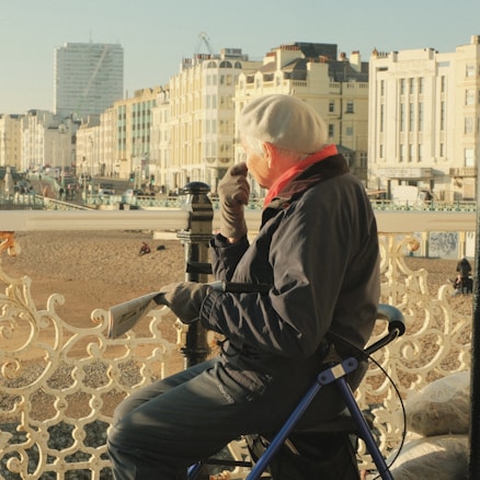 An elderly person wearing a white beret, pink scarf, and gloves is seated on a portable chair by a seaside promenade. They are looking at the beach and distant buildings, with a newspaper in hand. The scene features Victorian-style railings and several classic buildings along a street, basked in warm sunlight.