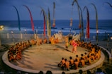 A traditional outdoor performance is underway near the ocean, featuring dancers in colorful costumes surrounded by a circle of seated spectators. The setting includes decorative swan statues, tall banners, and torchlights, creating a vibrant and cultural atmosphere.