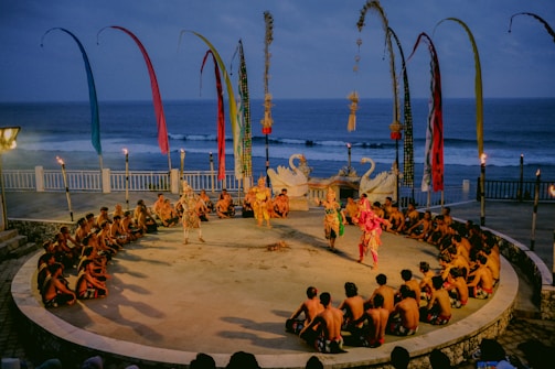A traditional outdoor performance is underway near the ocean, featuring dancers in colorful costumes surrounded by a circle of seated spectators. The setting includes decorative swan statues, tall banners, and torchlights, creating a vibrant and cultural atmosphere.