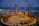 A traditional outdoor performance is underway near the ocean, featuring dancers in colorful costumes surrounded by a circle of seated spectators. The setting includes decorative swan statues, tall banners, and torchlights, creating a vibrant and cultural atmosphere.