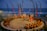 A traditional outdoor performance is underway near the ocean, featuring dancers in colorful costumes surrounded by a circle of seated spectators. The setting includes decorative swan statues, tall banners, and torchlights, creating a vibrant and cultural atmosphere.