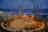 A traditional outdoor performance is underway near the ocean, featuring dancers in colorful costumes surrounded by a circle of seated spectators. The setting includes decorative swan statues, tall banners, and torchlights, creating a vibrant and cultural atmosphere.