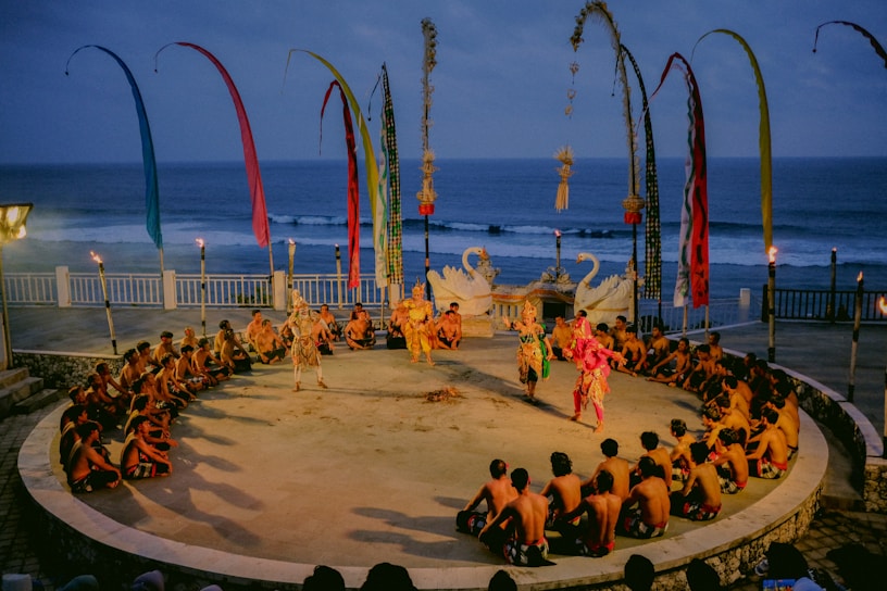A traditional outdoor performance is underway near the ocean, featuring dancers in colorful costumes surrounded by a circle of seated spectators. The setting includes decorative swan statues, tall banners, and torchlights, creating a vibrant and cultural atmosphere.