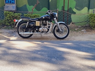 A classic Royal Enfield motorcycle is parked on the side of a road against a backdrop of green painted walls and foliage. The motorcycle has a distinct vintage look with chrome details and a black seat.