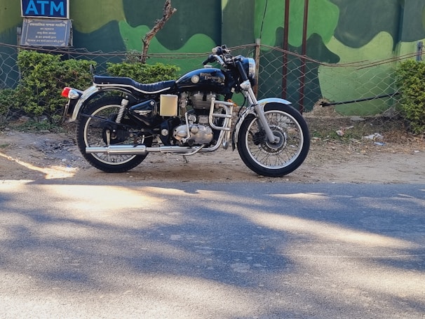 A classic Royal Enfield motorcycle is parked on the side of a road against a backdrop of green painted walls and foliage. The motorcycle has a distinct vintage look with chrome details and a black seat.
