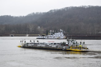 A river scene featuring a barge carrying several vehicles, likely cars, on a wide, calm river. In the background, a tugboat or larger vessel is visible with hills and trees lining the horizon under an overcast sky.