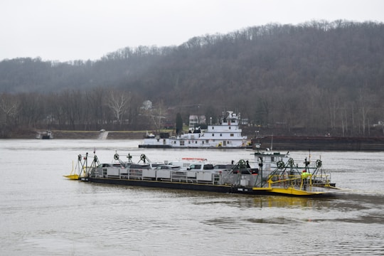 A river scene featuring a barge carrying several vehicles, likely cars, on a wide, calm river. In the background, a tugboat or larger vessel is visible with hills and trees lining the horizon under an overcast sky.