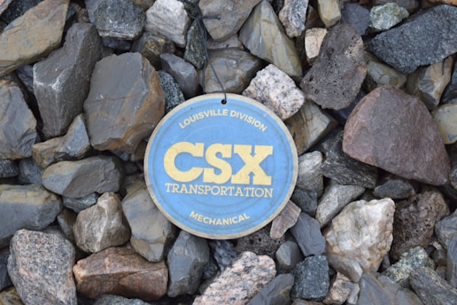 A circular sign with the text 'CSX Transportation' along with 'Louisville Division' and 'Mechanical' is placed on a background of variously sized and colored rocks.