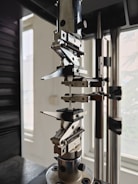 A close-up view of a mechanical testing apparatus used for material analysis, featuring metal clamps and screws against a background with windows.