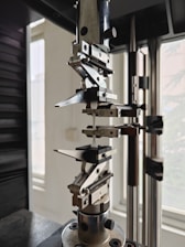 A close-up view of a mechanical testing apparatus used for material analysis, featuring metal clamps and screws against a background with windows.