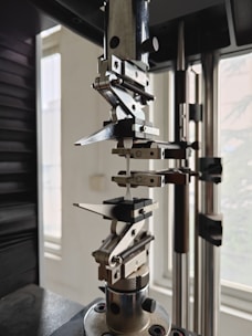A close-up view of a mechanical testing apparatus used for material analysis, featuring metal clamps and screws against a background with windows.