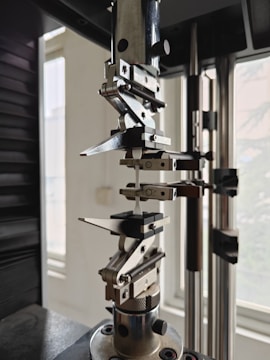 A close-up view of a mechanical testing apparatus used for material analysis, featuring metal clamps and screws against a background with windows.