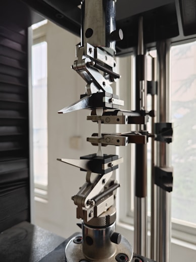 A close-up view of a mechanical testing apparatus used for material analysis, featuring metal clamps and screws against a background with windows.