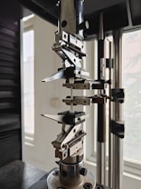 A close-up view of a mechanical testing apparatus used for material analysis, featuring metal clamps and screws against a background with windows.