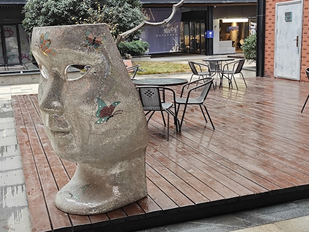 A large, textured sculpture of a face with butterfly designs stands on a wooden deck. There are several empty metal chairs and tables scattered around. In the background, a modern building with glass windows is visible, along with some greenery.