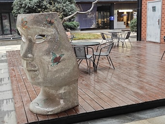 A large, textured sculpture of a face with butterfly designs stands on a wooden deck. There are several empty metal chairs and tables scattered around. In the background, a modern building with glass windows is visible, along with some greenery.