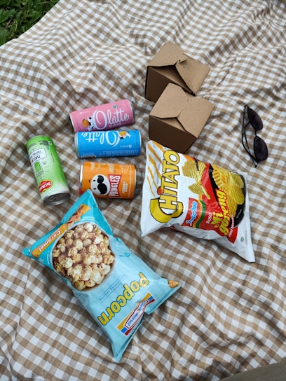 A picnic setup on a checkered blanket featuring several snack items. There are bags of potato chips and popcorn, different cans of drinks, two closed cardboard food boxes, and a pair of sunglasses. The surroundings suggest an outdoor setting, possibly in a park.