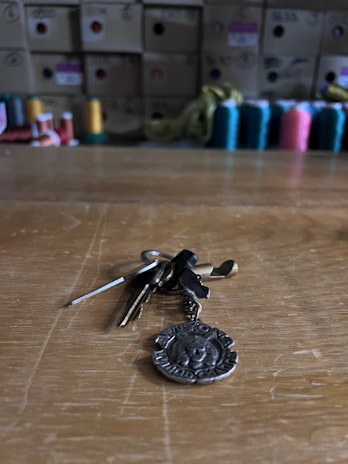 A set of keys with a metallic keychain lying on a wooden surface. In the background, there are colorful spools of thread and cardboard boxes stacked on shelves.