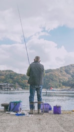 A person is fishing by a waterfront, sitting on a stool with scenic hills in the background. The setup includes various containers and equipment nearby, indicating preparation for a fishing activity.