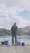 A person is fishing by a waterfront, sitting on a stool with scenic hills in the background. The setup includes various containers and equipment nearby, indicating preparation for a fishing activity.