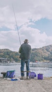 A person is fishing by a waterfront, sitting on a stool with scenic hills in the background. The setup includes various containers and equipment nearby, indicating preparation for a fishing activity.