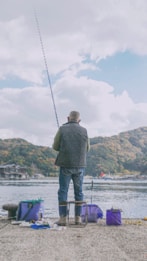 A person is fishing by a waterfront, sitting on a stool with scenic hills in the background. The setup includes various containers and equipment nearby, indicating preparation for a fishing activity.