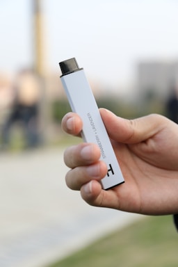 A hand is holding a rectangular vaping device with the words 'strawberry lavender' printed on its side. The device is white with a black mouthpiece. The background appears blurred, suggesting the image is focused on the vaping device in the foreground.