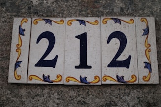 A set of decorative tiles displaying the number 212 against a textured stone background. The tiles feature ornate floral patterns in yellow and blue around the numbers.