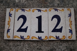 A set of decorative tiles displaying the number 212 against a textured stone background. The tiles feature ornate floral patterns in yellow and blue around the numbers.