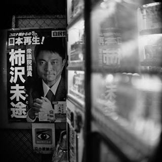 A black and white photograph featuring a political poster with Japanese text on it, displayed next to a vending machine. The poster depicts a man in a suit. The foreground includes the side of the vending machine with items visible through its glass.