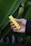 A hand is holding a yellow vape pen against a background of large green leaves. The pen has black accents and some text printed on it.