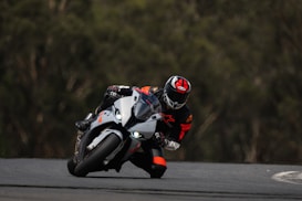 A motorcyclist in full racing gear, including a helmet and a leather suit, is leaning into a sharp curve on a racetrack. The motorcycle is a sleek, modern design and the rider is focused and in control, with bright headlights turned on. The background is a blur of green, suggesting speed and motion.