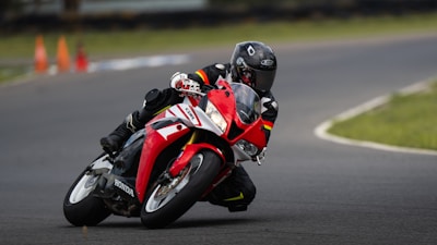A motorcyclist is leaning into a sharp turn on a racing track, riding a red and white sports motorcycle. The rider is dressed in full protective gear, including a helmet and a black suit with colorful stripes. There are orange traffic cones visible in the background, blurred due to motion.