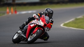 A motorcyclist is leaning into a sharp turn on a racing track, riding a red and white sports motorcycle. The rider is dressed in full protective gear, including a helmet and a black suit with colorful stripes. There are orange traffic cones visible in the background, blurred due to motion.
