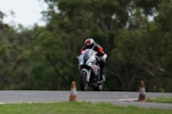 A motorcyclist wearing protective gear, including a helmet and a suit, is riding a white and black sport motorcycle on a track. The bike is slightly airborne, indicating high speed or a jump. The background consists of blurred green trees, and there are traffic cones near the track.