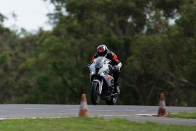 A motorcyclist wearing protective gear, including a helmet and a suit, is riding a white and black sport motorcycle on a track. The bike is slightly airborne, indicating high speed or a jump. The background consists of blurred green trees, and there are traffic cones near the track.